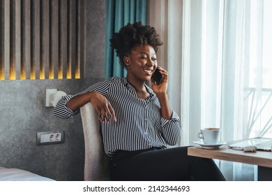 A Happy Business Woman Is Talking On A Mobile Phone While Sitting At The Table Of A Hotel Room With A Laptop. A Female CEO On A Business Trip Works From A Hotel Room.