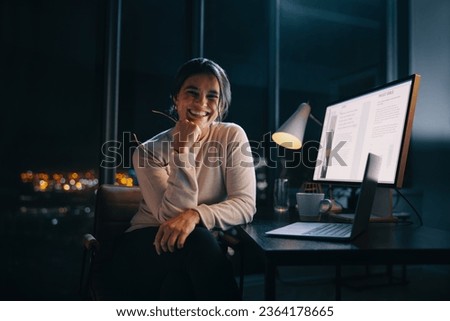 Happy business woman works late with her laptop and computer in a nighttime office. She plans and executes content marketing strategies after hours, showcasing her dedication and creativity.