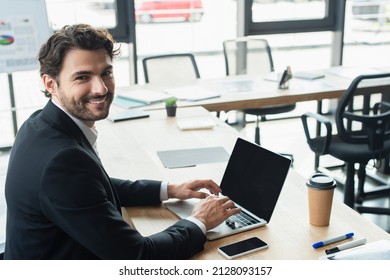happy businessman in black blazer typing on laptop and looking at camera in office