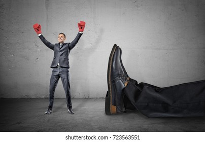 A happy businessman with boxing gloves on arms raised in victory stands near a giant male leg fallen down. Fight off competition. Unexpected winner. Chance of success.