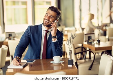 Happy businessman communicating over mobile phone and writing in notebook while sitting in a cafe.  - Powered by Shutterstock