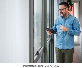 Happy Businessman Discussing While Video Conferencing On Digital Tablet. Male Executive Talking Through Wireless Computer. He Is Standing By The Window At Corporate Office.