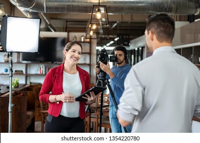 Happy businessman during corporate interview with female journalist. Manager answering question in office. Young woman at work as reporter with business man and cameraman shooting video for broadcast