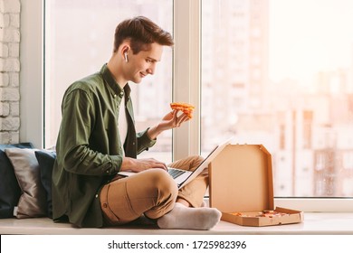 Happy businessman eat slice of tasty pizza, work remotely from home. Young hipster man freelancer sit on windowsill, using laptop for distance education, work, study, eat italian takeaway fast food - Powered by Shutterstock