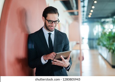 Happy Businessman In Formal Wear Standing Against The Wall In Business Center And Using Tablet For Reading E-mail.