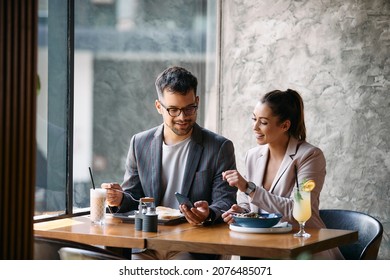 Happy businessman and his female colleague using mobile phone while eating breakfast in a cafe.