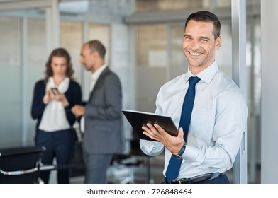 Happy Businessman Holding Digital Tablet With Team Discussing Project In The Background. Portrait Of A Confident Business Man Holding Computer Laptop. Successful Young Business Man At The Office. 
