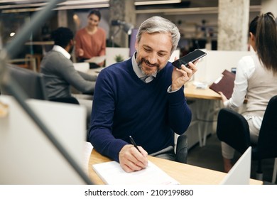 Happy Businessman Listening Voicemail On Smart Phone And Writing Notes While Working At Corporate Office. 