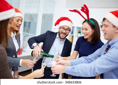 Happy businessman in Santa cap pouring champagne into flutes of his colleagues in office