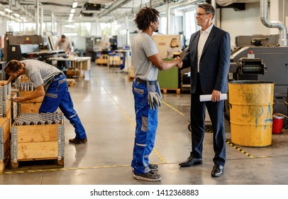 Happy businessman shaking hands with African American metal worker in industrial facility. 