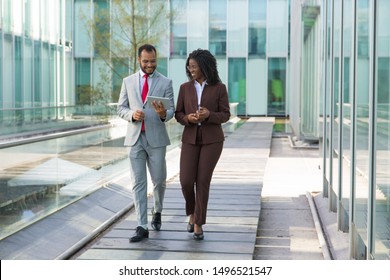 Happy Businessman Showing Content On Tablet To Female Colleague. Business Man And Woman Walking Outside, Using Tablet, Talking, Smiling, Laughing. Communication Concept