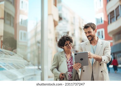 Happy businessman showing content on tablet to female colleague. Business man and woman walking outside, using tablet, talking, smiling, laughing. Communication concept - Powered by Shutterstock