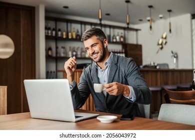 Ein glücklicher Geschäftsmann sitzt in einem Café, trinkt Kaffee und sieht sich ein Webinar auf dem Laptop an. Der Mann hat Ohrhörer, damit er den Vortrag hören kann. Ein Geschäftsmann, der im Café einen Laptop benutzt