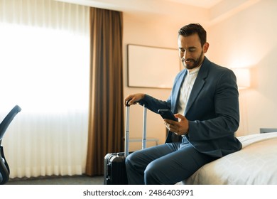 Happy businessman sitting on a bed and using a smartphone while holding a suitcase after arriving at a hotel room - Powered by Shutterstock