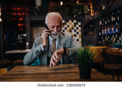 Happy Businessman Sitting In Restaurant And Waiting For Lunch. He Is Using Smart Phone And Talking With Someone. Business Seniors Lifestyle Concept.