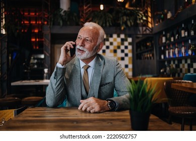 Happy Businessman Sitting In Restaurant And Waiting For Lunch. He Is Using Smart Phone And Talking With Someone. Business Seniors Lifestyle Concept.