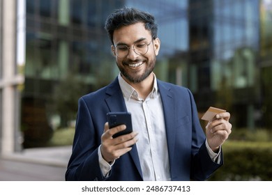 Happy businessman standing outdoors holding smartphone and credit card, making mobile payment. Professional wearing formal suit smiling while using technology for online transaction. - Powered by Shutterstock