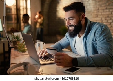 Happy businessman using laptop and credit card for online banking while working at night in the office.  - Shutterstock ID 1613789128