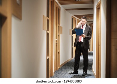 Happy businessman working in office corridor, financial consultant talking on mobile telephone and smiling. Adult bank manager standing and speaking with client on smartphone. - Shutterstock ID 1294803883
