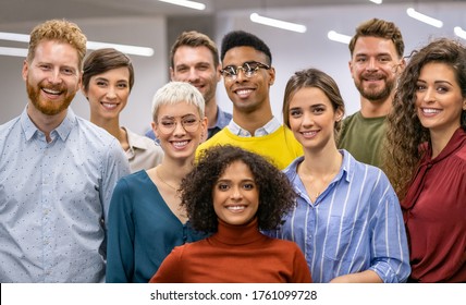 Happy businessmen and satisfied businesswomen standing together at creative office and looking at camera. Portrait of successful multiethnic group of casual men and women smiling at modern workplace.
