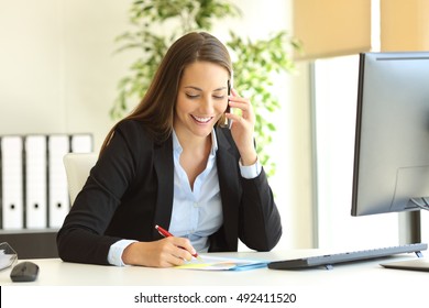 Happy businesswoman calling on mobile phone and taking notes on a desk at office