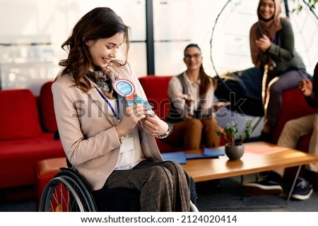 Happy businesswoman with disability receiving employee of the month badge while working at corporate office. Group of colleagues are applauding her in the background.