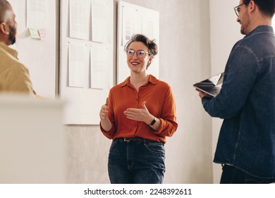 Happy businesswoman having a discussion with her team next to some notice boards. Group of cheerful business colleagues sharing ideas during a meeting in a creative office. - Powered by Shutterstock