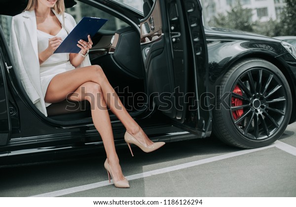 Happy businesswoman\
making notes in document while locating in contemporary vehicle\
with opened door. She wearing attractive shoes and throwing leg on\
another one
