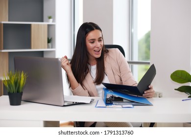 Happy Businesswoman With Raised So Hand Gesture Reading Letter On Desk In Front Of Laptop. The Businessman Is Satisfied With The Good News From The Correspondence.