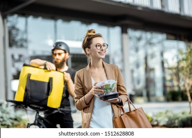 Happy Businesswoman Receiving Fresh Takaway Lunches, Standing Near The Office Building With A Male Courier On The Background. Takeaway Restaurant Food Delivery Concept