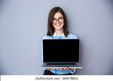 Happy Businesswoman Showing Blank Laptop Screen Over Gray Background. Wearing In Blue Shirt And Glasses. Looking At Camera