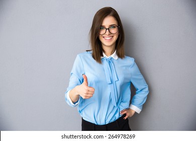 Happy businesswoman showing thumb up over gray background. Wearing in blue shirt and glasses. Looking at camera