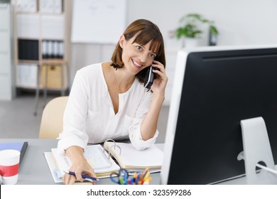 Happy Businesswoman Sitting At Her Desk, Talking To Someone On Mobile Phone While Working On Her Computer