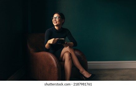 Happy businesswoman sitting on armchair with cup of tea. Beautiful woman relaxing on armchair and looking away. - Powered by Shutterstock