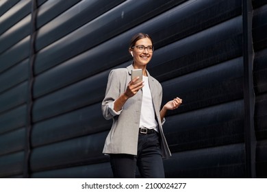 A happy businesswoman walking outdoors and having phone calls over earbuds. Busy woman making choices on a way to her office. A businesswoman with a phone and earphones - Shutterstock ID 2100024277