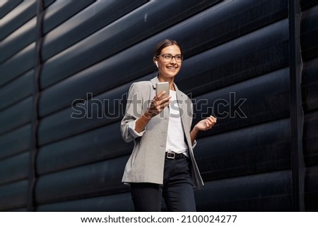 A happy businesswoman walking outdoors and having phone calls over earbuds. Busy woman making choices on a way to her office. A businesswoman with a phone and earphones