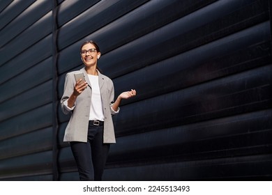 A happy businesswoman walking outdoors and listening to a podcast over her phone. A businesswoman educating herself while walking. A businesswoman with phone - Powered by Shutterstock