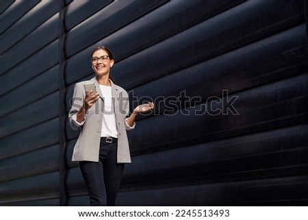 A happy businesswoman walking outdoors and listening to a podcast over her phone. A businesswoman educating herself while walking. A businesswoman with phone