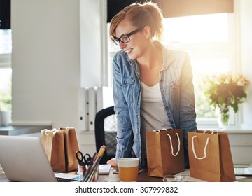 Happy Businesswoman Working In A Home Office Standing Looking At Her Laptop Computer Surrounded By Gift Bags And Packaging For The Products For Sale On Her New Website