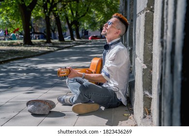 Happy Busker Street Musician Sitting On A City Sidewalk And Playing Music With Guitar. Freedom, Music And Art Concept.
