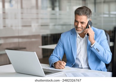 Happy busy 50 years old business man talking with client on mobile phone working on laptop using computer in office. Smiling mature senior businessman entrepreneur making call on cellphone at work. - Powered by Shutterstock