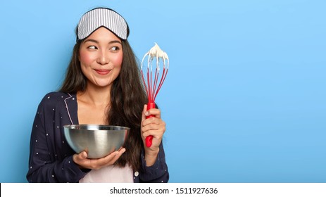 Happy Busy Asian Housewife Whisks Cream In Bowl, Wears Sleep Mask And Pyjamas, Cooks Early In Morning After Aweking, Poses Against Blue Background. Woman Makes Homemade Cake. Cooking Concept