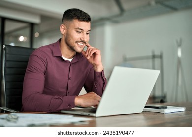 Happy busy Latin business man employee or manager working on digital technology project sitting at desk. Young smiling professional businessman entrepreneur using computer laptop in office.