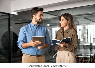 Happy busy latin partners using digital tablet talking standing in office. Professional business people having conversation sharing ideas. Female executive manager working together with male employee. - Powered by Shutterstock