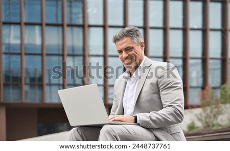 Happy busy middle aged business man working on laptop outside office. Smiling older professional businessman executive ceo manager wearing suit sitting outdoors using computer managing data.