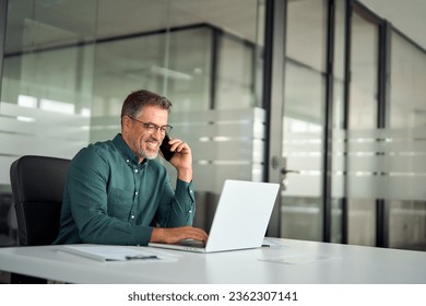 Happy busy older mid aged business man professional expert or entrepreneur making phone call speaking with client communicating on cellphone using laptop computer sitting at desk in office. Copy space - Powered by Shutterstock