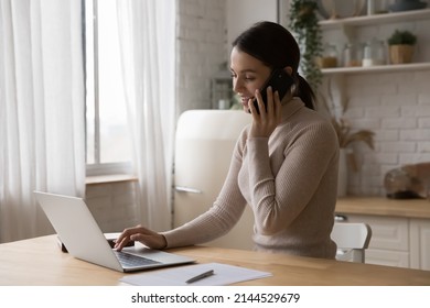 Happy Busy Young Freelancer Woman Working In Home Kitchen, Speaking On Mobile Phone, Using Laptop Computer At Table, Laughing, Giving Consultation. Remote Job, Communication Concept