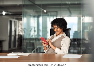 Happy busy young professional African American business woman executive holding cellphone using mobile looking at mobile cell phone tech with smartphone in hands sitting at desk in office working. - Powered by Shutterstock