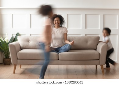 Happy Calm African American Mom Sit On Couch Relax Practicing Yoga Distracted From Active Kids Running Around, Smiling Black Mother Meditate On Sofa With Noisy Children Having Fun. Stress Free Concept