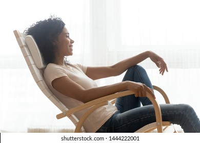Happy calm african girl resting dreaming sit in comfortable rocking chair relaxing on cozy weekend, smiling black woman lounging enjoy peaceful quiet lifestyle feeling no stress free at comfy home